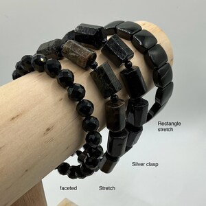 May include: Three black gemstone bracelets displayed on a wooden stand. One bracelet features faceted beads, another has rectangular beads, and the third has a stretch design. The bracelets are a dark, glossy black.