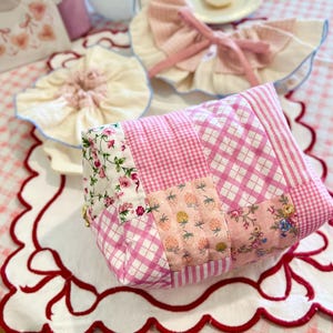 以下が含まれることがあります： ピンクのギンガムチェックと花柄のパッチワークバッグ。赤い縁取りのある白いスカラップのプレースマットの上にバッグがあり、フリル付きのよだれかけやクッキーの入った小さなプレートなどの装飾品が並んでいます。