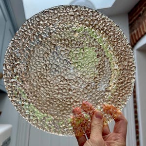Puede incluir: Un plato de vidrio transparente con un patrón texturizado y burbujeado. El plato se sostiene, mostrando su intrincado diseño y refracción de la luz. Los bordes del plato son ligeramente irregulares.