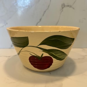 Puede incluir: Cuenco de cerámica color crema con un diseño pintado a mano de una manzana roja y hojas verdes. El cuenco tiene una forma ligeramente acampanada y una estética rústica y vintage. El diseño de la manzana y la hoja se repite.