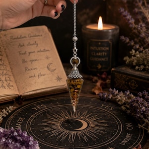 May include: A pointed, cone-shaped pendulum filled with small, golden-brown metallic pieces, topped with an ornate silver-toned metal cap and a delicate silver chain. The pendulum hangs vertically, featuring a clear, transparent casing.