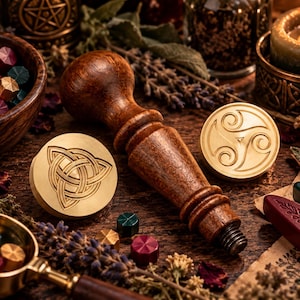 May include: A dark brown wooden handle for a wax seal stamp, accompanied by two circular brass stamp heads. One head features an engraved triquetra knot design, while the other displays a triskelion spiral pattern. Small wax beads are scattered nearby.