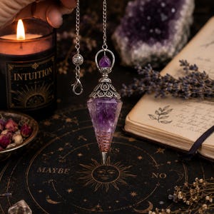 May include: A faceted purple amethyst pendulum with a silver-toned metal cap featuring intricate filigree detailing. The crystal is wrapped in copper wire at the pointed base and hangs from a silver-colored chain with a small lobster clasp.