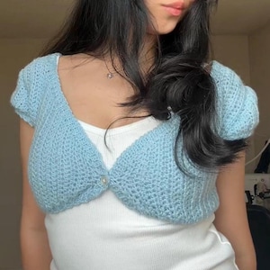May include: A light blue, short-sleeved, cropped crochet cardigan featuring a single button closure at the center front. The garment is worn over a plain white tank top, showcasing a textured knit pattern and a V-neckline design.