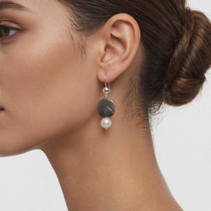 May include: A drop earring featuring a silver hook, a small silver bead, a flat circular dark grey stone disc with subtle veining, and a single round white pearl at the base. The earring hangs vertically from the earlobe with a polished finish.