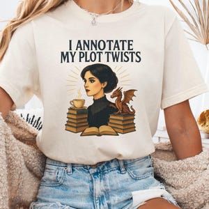 Puede incluir: Camiseta color crema con el texto "I Annotate My Plot Twists". El gráfico presenta una mujer, un dragón, una taza de café y pilas de libros. El diseño es de estilo vintage.