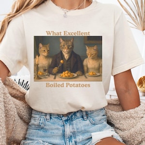 May include: Cream-colored t-shirt featuring a humorous image of three cats at a table, dressed in period clothing, with the text "What Excellent Boiled Potatoes." The cats are depicted in a parody of a classic painting.