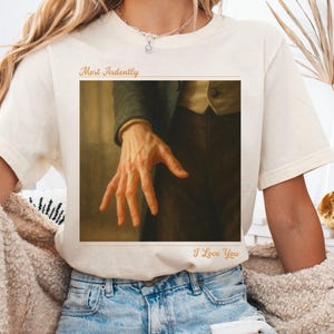 May include: Off-white t-shirt with a framed image of a hand reaching out, with the words "Most Ardently" and "I Love You" in gold. The shirt is worn with blue jeans.