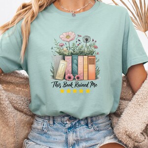 May include: A pale blue t-shirt featuring a design of books and flowers, with the text "This Book Ruined Me" and five yellow stars. The graphic includes pink, orange, and green colours. The t-shirt is a relaxed fit and suitable for everyday wear.