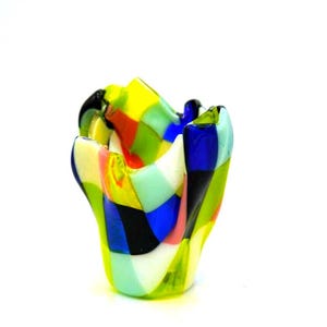 May include: A colorful, handcrafted glass vase with a unique, organic shape. The vase features a patchwork design of vibrant colors including yellow, green, blue, black, and pink. The vase is a decorative item.