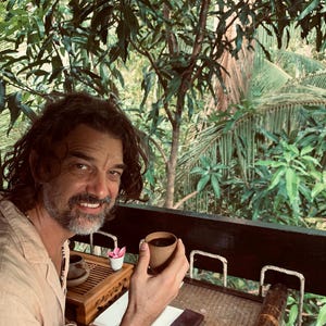 Può includere: Una persona sorridente che tiene in mano una piccola tazza marrone di tè, con un lussureggiante sfondo verde di fogliame tropicale. Un set da tè in legno, un taccuino e un piccolo oggetto incorniciato sono sul tavolo. La persona indossa una camicia di colore chiaro.