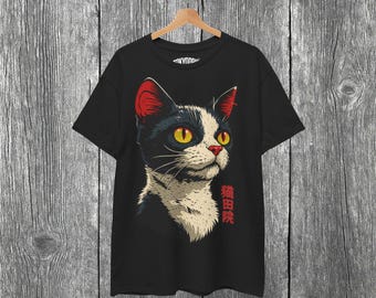 May include: Black t-shirt featuring a stylized cat portrait. The cat has black fur with white markings, red ears, and yellow eyes. Japanese text is printed vertically on the right side of the cat. The shirt is hanging on a wooden hanger against a gray wood-paneled background.