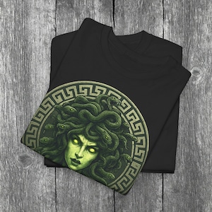Könnte beinhalten: Schwarzes T-Shirt mit einer Grafik von Medusa, dargestellt mit grünen Schlangen als Haare und leuchtend gelben Augen. Das Design befindet sich in einem kreisförmigen Rahmen mit einem griechischen Schlüsselmuster. Das T-Shirt ist auf einer Holzoberfläche gefaltet.