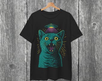 May include: Black t-shirt featuring a teal cat with wide yellow eyes and an open mouth, under a purple and silver UFO. The cat has a surprised expression, with yellow lines radiating outwards.