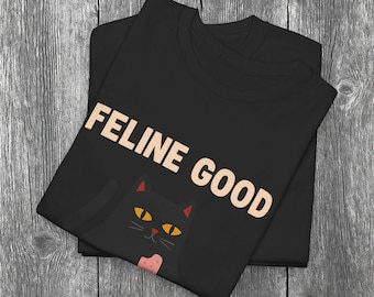Pode incluir: Camiseta preta com o texto "FELINE GOOD" em castanho claro. Um gato preto de desenho animado com olhos amarelos e uma gravata borboleta rosa é apresentado abaixo do texto. A camiseta está dobrada sobre uma superfície de madeira.