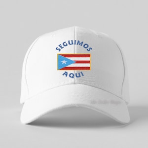 Puede incluir: Gorra de béisbol blanca con la bandera de Puerto Rico y las palabras "SEGUIMOS AQUI" en azul. El diseño de la bandera incluye un triángulo azul con una estrella blanca y rayas rojas y blancas.