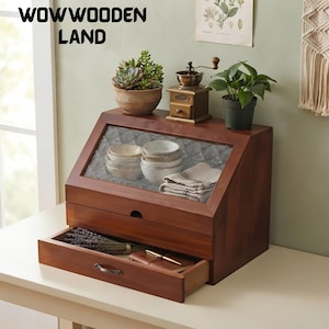 May include: A wooden bread box with a slanted glass front panel, a top surface holding a potted succulent, a small coffee grinder, and a green plant, and a bottom drawer containing a bundle of dried lavender and a small notebook with a gold pen.