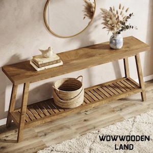 May include: A long wooden console table with a rectangular top and a slatted lower shelf. On the table sits a stack of three books topped with a small bird figurine, and a blue and white vase filled with dried pampas grass and eucalyptus stems.