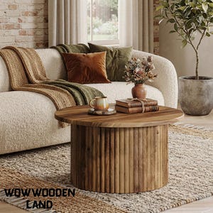 May include: A round wooden coffee table with a fluted, cylindrical base and a smooth, circular top. The table is crafted from natural wood with visible grain patterns, featuring a sturdy, solid construction that sits directly on the floor.