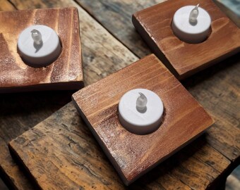 May include: Three square wooden candle holders with a warm brown finish. Each holder has a circular indentation holding a white LED tea light. The holders are arranged on a rustic wooden surface.