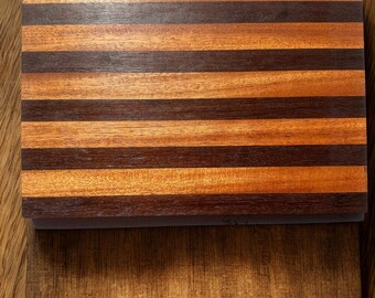 May include: A rectangular wooden cutting board with alternating dark brown and light brown stripes. The board is made of hardwood and has a smooth, polished finish. The cutting board is shown on a wooden surface.