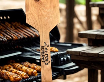 May include: A wooden grill scraper with the text "Lauren's scraper" in black script. The scraper is light brown and has a knot in the wood. The background shows a grill with food and a wooden picnic table.