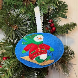 May include: A blue felt Christmas ornament featuring a snowman skiing on a wooden sled. The snowman wears a red coat, green scarf, and green mittens. The ornament is oval-shaped and has a white ribbon for hanging.