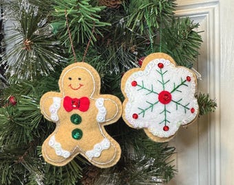 May include: Two felt Christmas ornaments hanging from a tree. One is a tan gingerbread person with a red bow tie, green sequin buttons, and white rickrack trim. The other is a white snowflake shape with green embroidery and red sequins.