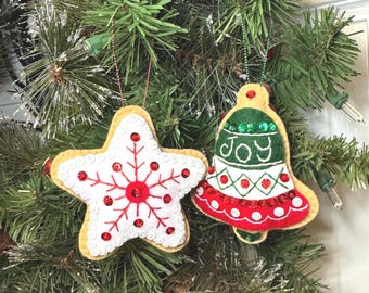 May include: Two felt Christmas ornaments hanging from a tree. One is a white star with red embroidered snowflake details and red sequins. The other is a bell-shaped ornament in green, white, and red felt with the word JOY embroidered in white.