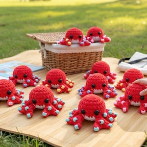 May include: A collection of red crocheted octopus toys with white faces and colorful tentacles are arranged on a wooden surface. A wicker basket and a child's hand are also visible. The scene is set outdoors on a picnic blanket.