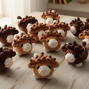 May include: A collection of small, round, crocheted highland cow figures. Each figure features a textured brown or tan yarn body, a white muzzle, small black bead eyes, and a tuft of curly yarn hair on top. The figures are arranged on a wooden surface.