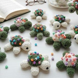 May include: A collection of handmade crochet turtles. Each turtle features a green shell adorned with colorful beads, white flipper-like appendages, and black eyes. The turtles are arranged on a white surface, suggesting a craft or toy theme.