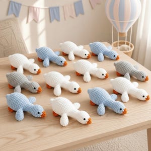 May include: A collection of eleven small, crocheted plush geese in white, light blue, and gray. Each goose features a textured yarn body, black bead eyes, and a small orange beak. The geese are arranged on a light wood surface in a group.