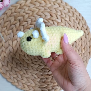 May include: A small, pale yellow crocheted triceratops dinosaur toy made from soft, plush yarn. The dinosaur features a white crocheted frill and horns, along with a single black plastic safety eye visible on the side of its rounded head.