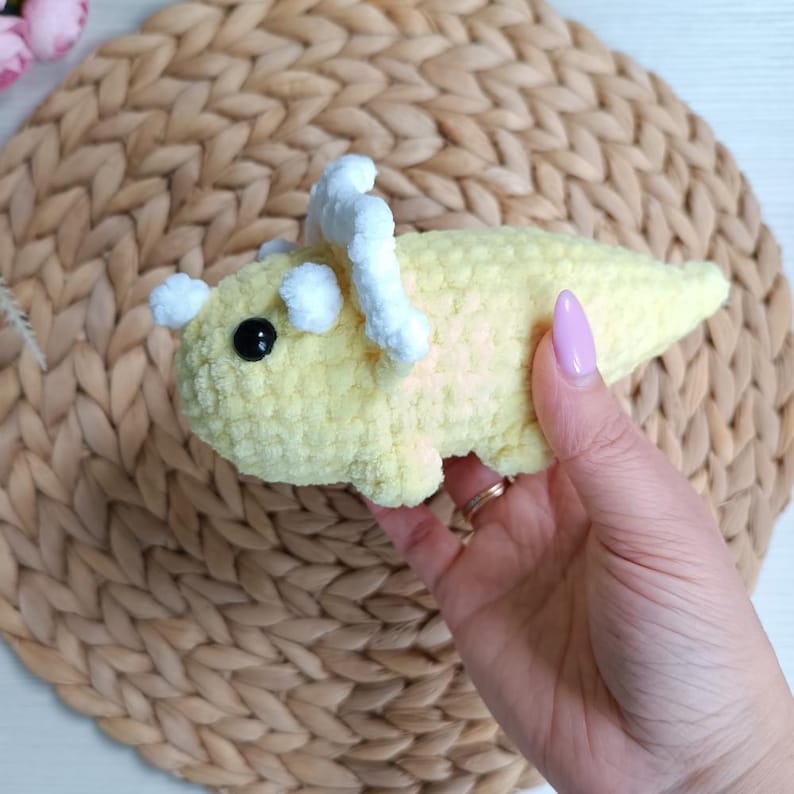 May include: A small, pale yellow crocheted triceratops dinosaur toy made from soft, plush yarn. The dinosaur features a white crocheted frill and horns, along with a single black plastic safety eye visible on the side of its rounded head.