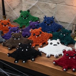 May include: A collection of small, crocheted plush creatures in various colors including green, orange, purple, blue, brown, black, white, and red. Each plush features two black plastic eyes, two small white fangs, and silver-toned beads at the tips.