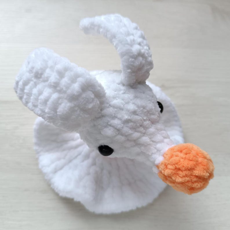 Puede incluir: Un juguete de peluche tejido a ganchillo en color blanco con forma de cabeza de fantasma, dos orejas erguidas, una nariz redonda naranja y peque&ntilde;os ojos de cuentas negras. La figura tiene una base de cuello con volantes de hilo blanco.