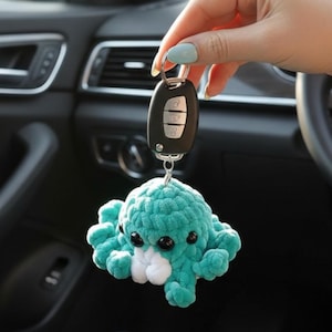 Peut inclure: Un porte-clés pieuvre en crochet bleu sarcelle avec de grands yeux noirs et un petit accent blanc près de la base, attaché à une clé de voiture en plastique noir avec trois boutons argentés. La pieuvre en peluche a une texture douce.