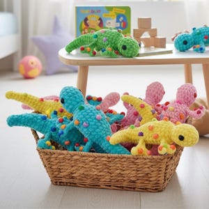 Può includere: Un cesto di vimini pieno di giocattoli di dinosauri all'uncinetto colorati. I dinosauri sono in tonalità di blu, giallo, rosa e verde, con macchie multicolori. Alcuni dinosauri sono su un tavolino con un libro e blocchi di legno.