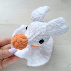 Puede incluir: Un juguete de peluche tejido a ganchillo en color blanco que representa un personaje fantasmag&oacute;rico con orejas grandes, una nariz naranja redonda y peque&ntilde;os ojos de bot&oacute;n negros. El hilo suave y texturizado forma una cabeza y una base fruncida.