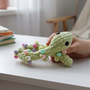 Puede incluir: Un juguete de serpiente de ganchillo verde claro hecho a mano con ojos de botón negros y detalles de cuentas de colores. La serpiente está sostenida en una mano. Las cuentas son de colores pastel. El juguete está sobre una superficie blanca.