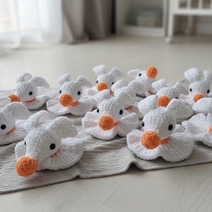 Puede incluir: Una colecci&oacute;n de peque&ntilde;as figuras de peluche tejidas a ganchillo en color blanco con forma de patos. Cada figura presenta un pico redondo y texturizado de color naranja, dos peque&ntilde;os ojos de cuentas negras y una base con volantes.