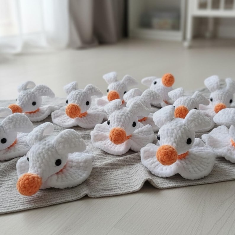 Puede incluir: Una colecci&oacute;n de peque&ntilde;as figuras de peluche tejidas a ganchillo en color blanco con forma de patos. Cada figura presenta un pico redondo y texturizado de color naranja, dos peque&ntilde;os ojos de cuentas negras y una base con volantes.