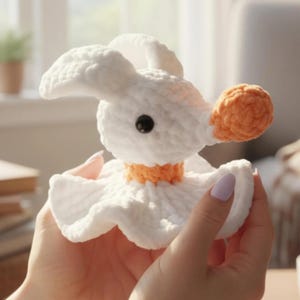Puede incluir: Un juguete de peluche tejido a ganchillo en color blanco con cabeza redondeada, orejas largas, un peque&ntilde;o ojo negro y una nariz naranja texturizada. La figura presenta un cuello naranja y una base blanca con volantes de hilo suave.