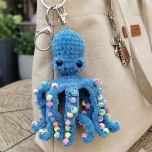 May include: A blue crocheted octopus keychain with black eyes and colorful beads on its tentacles. The plush octopus is attached to a silver keyring and is hanging on a beige tote bag. The octopus is a cute and whimsical accessory.