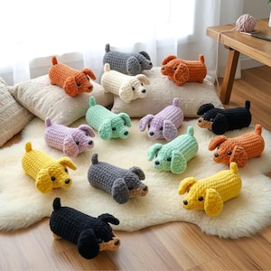 Könnte beinhalten: Eine Sammlung von zwölf kleinen, gehäkelten Dackel-Spielzeugen in verschiedenen Unifarben wie Gelb, Orange, Schwarz, Grau, Lavendel, Minzgrün und Creme. Jeder Plüschhund hat einen zylindrischen Körper, Schlappohren und kleine schwarze Augen.