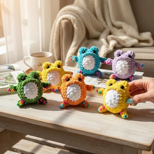 Puede incluir: Seis ranas de crochet hechas a mano en varios colores: verde, amarillo, naranja, azul, morado y amarillo. Cada rana tiene una barriga blanca y pequeños brazos y piernas con detalles coloridos. Las ranas se exhiben sobre una superficie de madera clara.