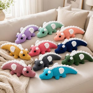 May include: A collection of nine small, crocheted plush crocodile toys in various solid colors including purple, mint green, peach, yellow, pink, dark blue, dark gray, dark green, and bright blue, each featuring white spikes and black eyes.