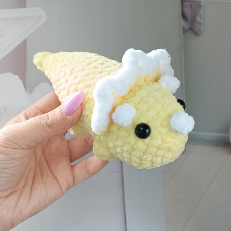 May include: A soft, crocheted triceratops plush toy in a pale yellow color. The dinosaur features a white frill and a small white horn on its snout. It has two round, black plastic eyes and a textured, yarn-based surface throughout its body.