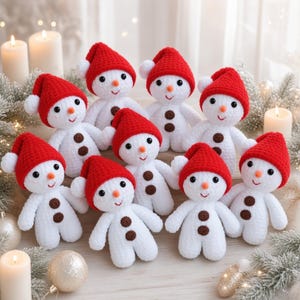 May include: A group of nine small, white crocheted snowman dolls, each wearing a bright red pointed hat with a white pom-pom. Each snowman features two brown circular buttons on its chest, a small orange nose, and black embroidered eyes and mouth.
